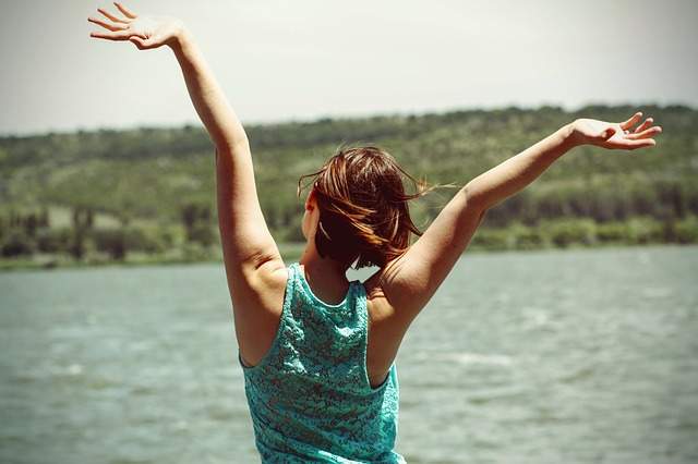 femme de dos portant un top sans manches et qui lève les bras debout devant un lac