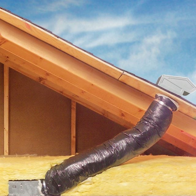 Une personne raccorde une gaine flexible isolée au conduit d'un chapeau de toiture dans un grenier.