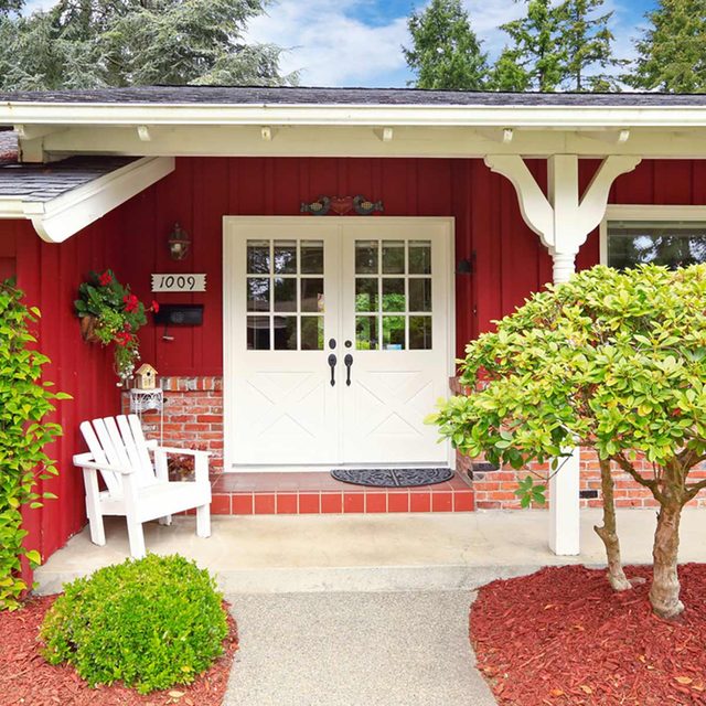 Une chaise blanche est placée sur le porche d'une maison rouge, à côté de la double porte d'entrée.