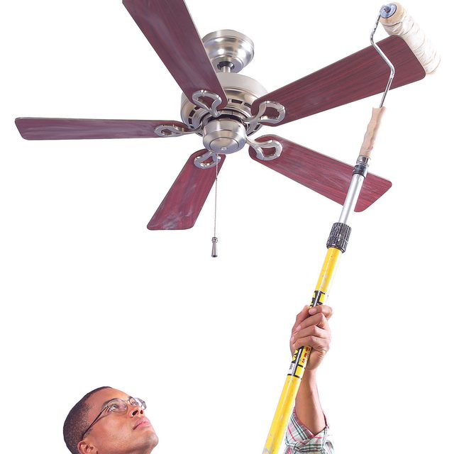 Une personne utilise un rouleau à peinture sur une perche pour dépoussiérer les pales d'un ventilateur de plafond.