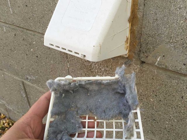 Une main retire un filtre blanc plein de peluches d'une bouche d'aération murale.