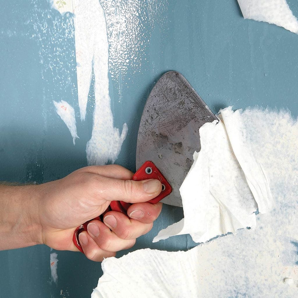 Une main décolle du papier peint d'un mur bleu à l'aide d'une spatule.