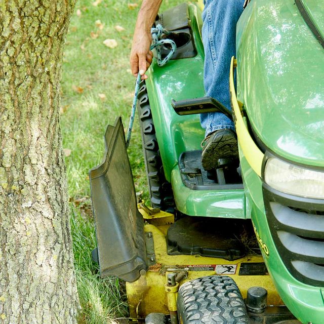 Une personne sur un tracteur tondeuse tirant sur une corde pour soulever le déflecteur d'herbe.