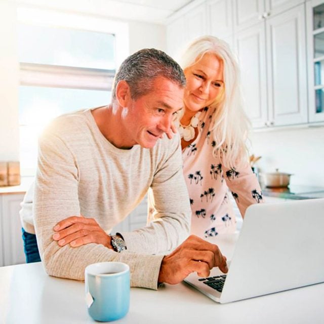 Un homme et une femme plus âgés sourient en regardant un ordinateur portable dans une cuisine.