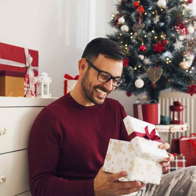 Un homme souriant ouvre un cadeau de Noël à côté d'un sapin décoré.