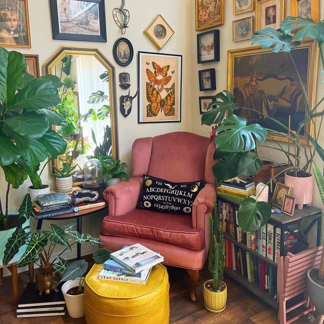 Un fauteuil rose avec un coussin décoratif, entouré de plantes, de livres et de cadres sur un mur.