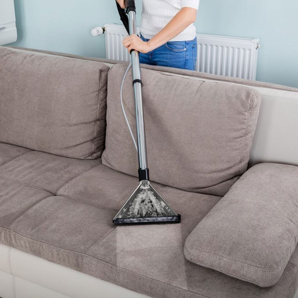 Une personne nettoie un canapé en tissu avec un nettoyeur vapeur.