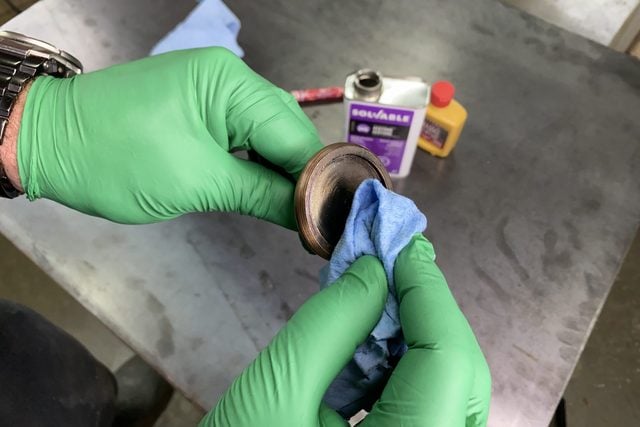 Une personne portant des gants verts essuie un objet métallique avec un chiffon bleu, entourée de produits de nettoyage.