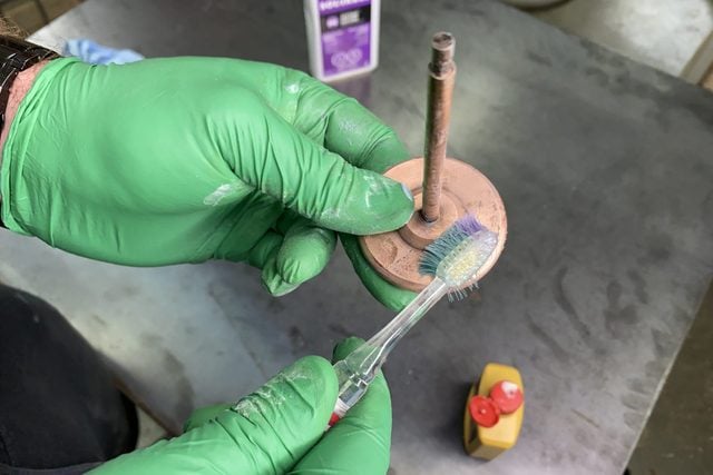 Une personne tient un objet en bois et le brosse avec une brosse à dents, à côté d'un flacon jaune.