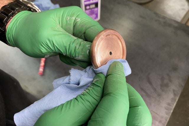 Une personne portant des gants verts nettoie un objet en cuivre avec un chiffon bleu.