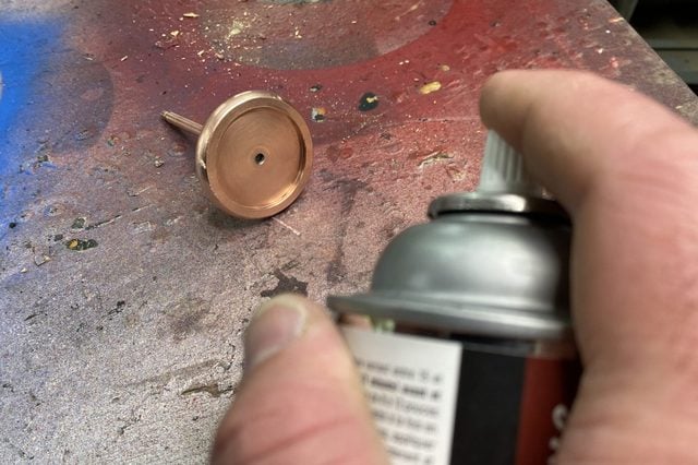 Une main pulvérise un produit sur un objet en cuivre posé sur un établi.