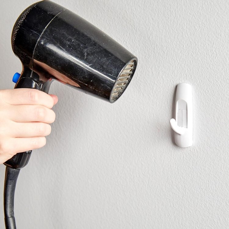 Un sèche-cheveux est pointé vers un crochet adhésif de type Command fixé sur un mur.