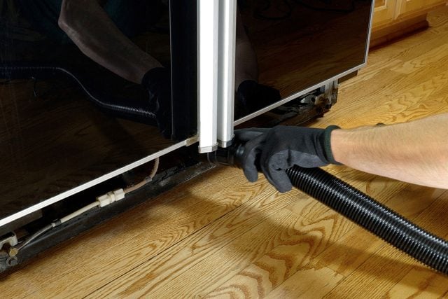 Une personne aspire la poussière de la grille d'un frigo avec un aspirateur de chantier.