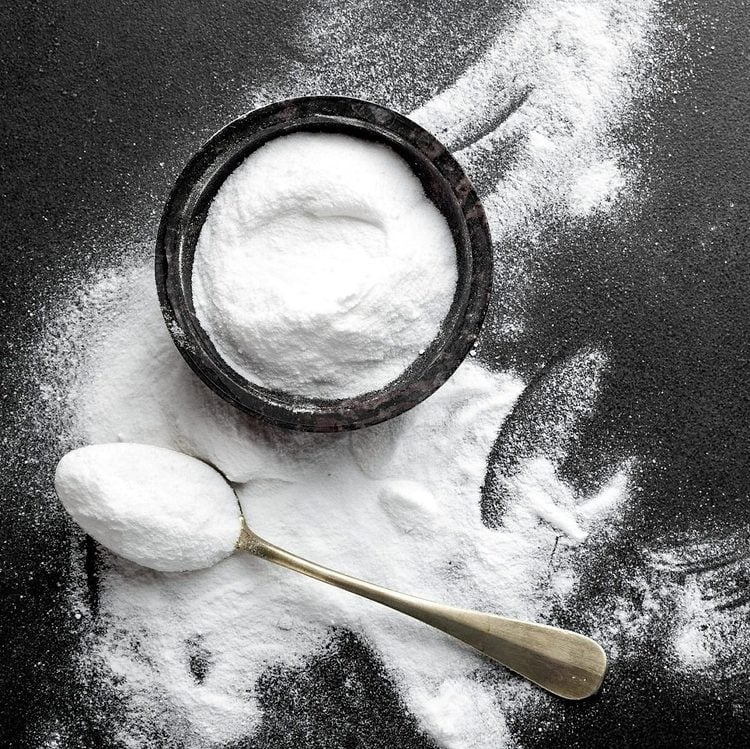 Un bol de poudre blanche sur une surface sombre, avec une cuillère de poudre à côté.