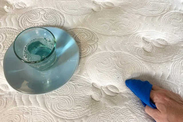 Une main utilisant un chiffon pour nettoyer une tache sur un matelas blanc.