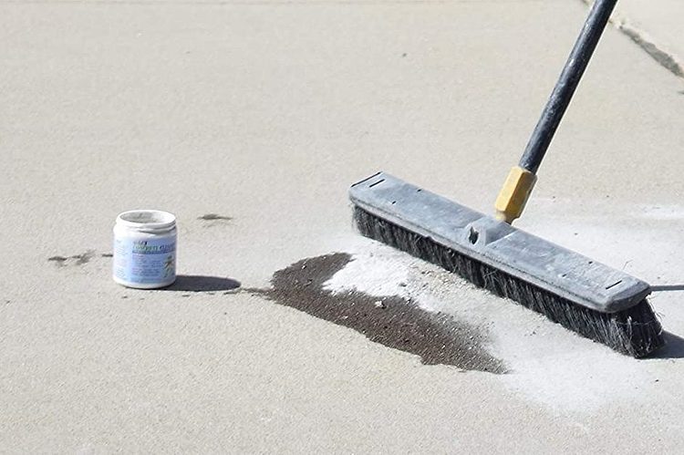 Un balai brosse nettoyant un sol en béton clair, avec un petit pot de produit nettoyant à proximité.