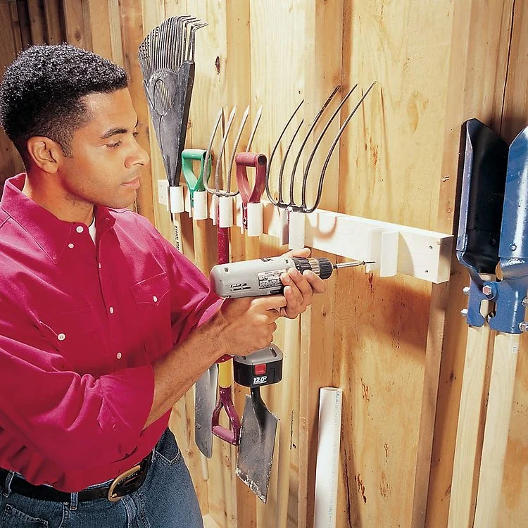 Un homme fixe une étagère en bois au mur d'un garage avec une perceuse.
